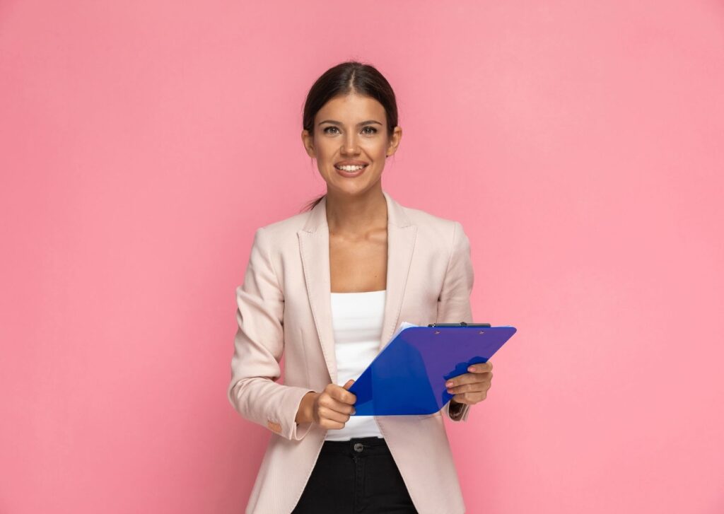 A white woman in a nude blazer holding a blue clipboard against a pink background.